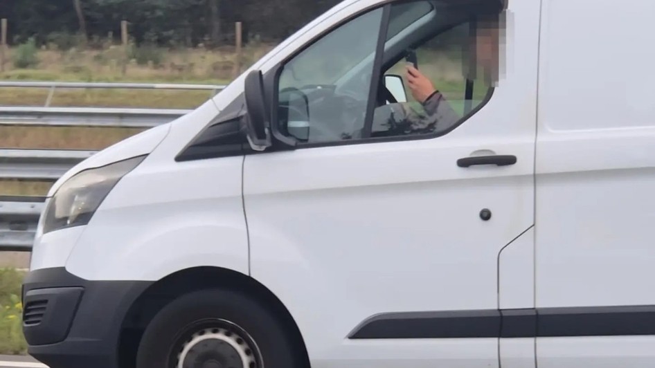 Imaginea prezintă un șofer (cu fața blurată) la volanul unei autoutilitare albe, utilizând un telefon mobil în timp ce conduce pe autostradă.
