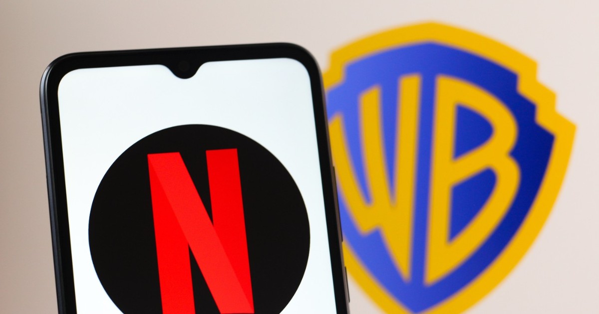 În imagine se vede un telefon pe al cărui ecran apare sigla Netflix, cu litera roșie „N” pe fundal negru. În spatele telefonului, neclar, se distinge logo-ul Warner Bros., sugerând contextul unui parteneriat sau al unei achiziții între cele două companii.