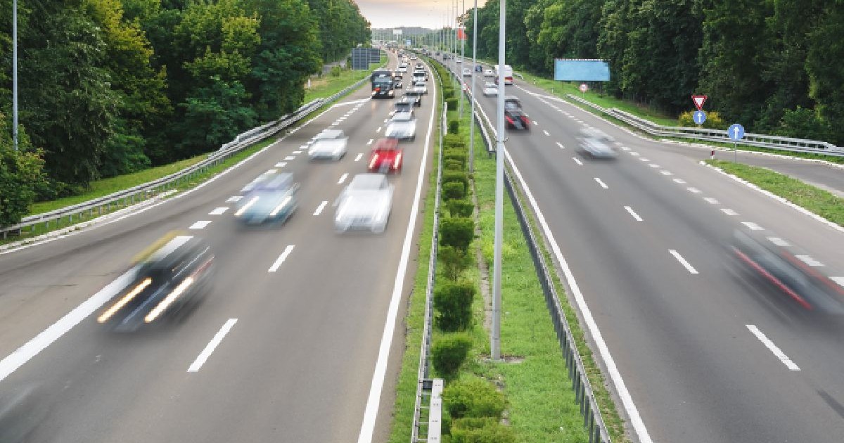 Fotografie cu o autostradă aglomerată, cu mai multe benzi, pe care circulă numeroase vehicule, inclusiv mașini și camioane, în ambele sensuri. Drumul este flancat de copaci verzi și mediană acoperită cu iarbă. Cerul este senin, cu o urmă de apus de soare