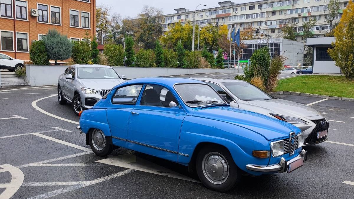 O mașină SAAB 96 albastră, staționată într-o parcare, lângă alte mașini moderne, în curtea RAR București Grivița