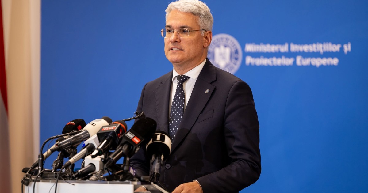 Imaginea surprinde un oficial care susține o declarație de presă la un pupitru, în fața mai multor microfoane ale instituțiilor media. În fundal se observă sigla și denumirea Ministerului Investițiilor și Proiectelor Europene, sugerând un eveniment oficial sau o conferință de presă.