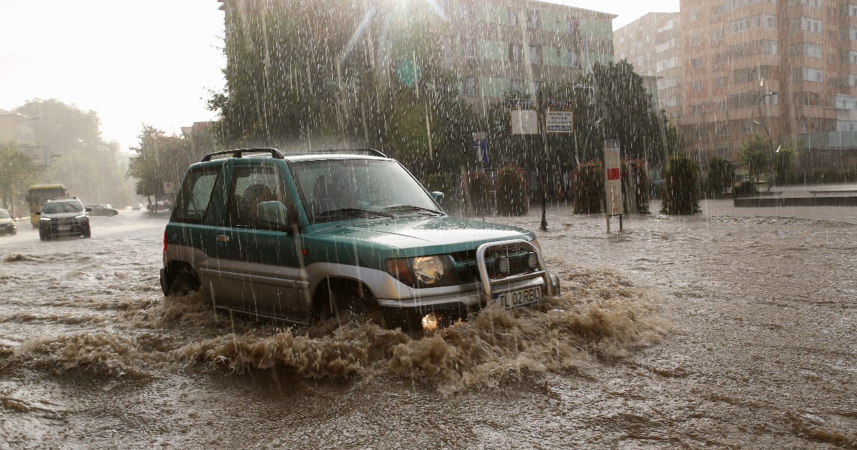 Imaginea surprinde un vehicul de teren (SUV) de culoare verde-turcoaz și argintiu care traversează o stradă complet inundată de apă maro, cel mai probabil în timpul unei ploi torențiale în Tulcea, România. Mașina creează valuri mari de apă murdară și este parțial acoperită, în timp ce pe fundal se văd clădiri de locuințe.