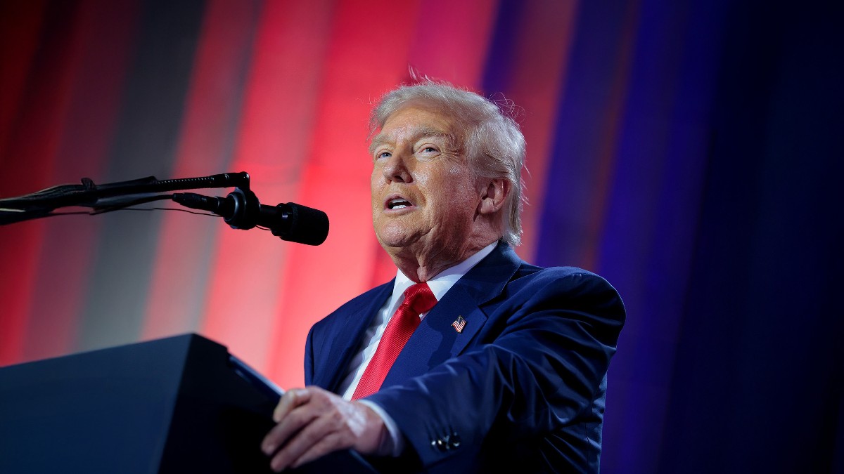 Imaginea îl prezintă pe Donald Trump, îmbrăcat într-un costum albastru închis cu o cravată roșie, vorbind la un microfon de la un pupitru în timpul unui discurs. În fundal se observă o scenă iluminată cu dungi de lumină roșie și albastră, care sugerează un eveniment public sau o adunare politică.