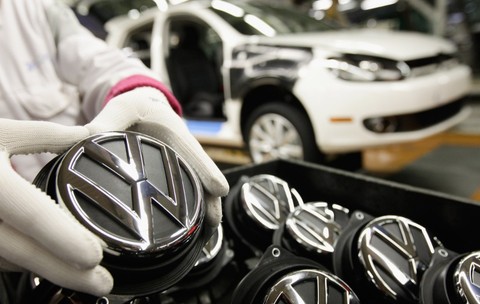 Imaginea prezintă un lucrător cu mănuși de protecție manevrând o emblemă cromatică Volkswagen pe linia de asamblare a unei fabrici auto.   