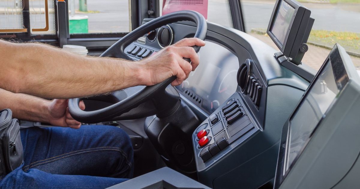 Un șofer este cu mâinile pe volanul unui autobuz și conduce pe o stradă