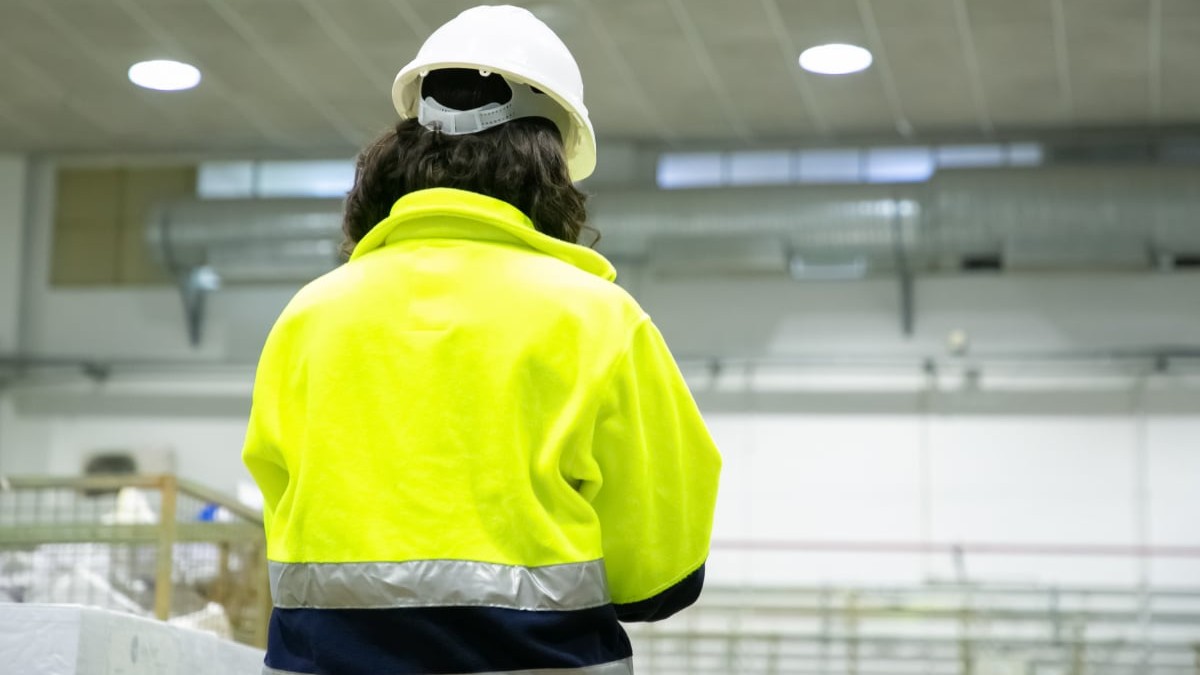 O muncitoare privită din spate poartă cască albă de protecție și o jachetă galben-fluorescentă reflectorizantă într-un spațiu industrial sau depozit.