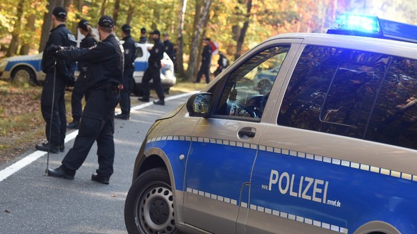 Mai mulți polițiști germani în uniforme desfășurând o operațiune de căutare sau securizare pe un drum de lângă o pădure, având o autospecială de poliție cu girofarul aprins în prim-plan.