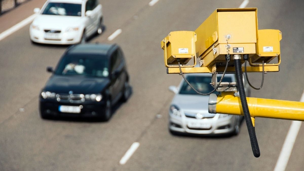 Vedere de sus a unei autostrăzi cu mai multe benzi. În prim-plan, un dispozitiv galben de monitorizare a traficului este montat pe un stâlp metalic. Trei mașini, un sedan negru, un hatchback argintiu și un sedan alb circulă în același sens, pe banzi diferite.