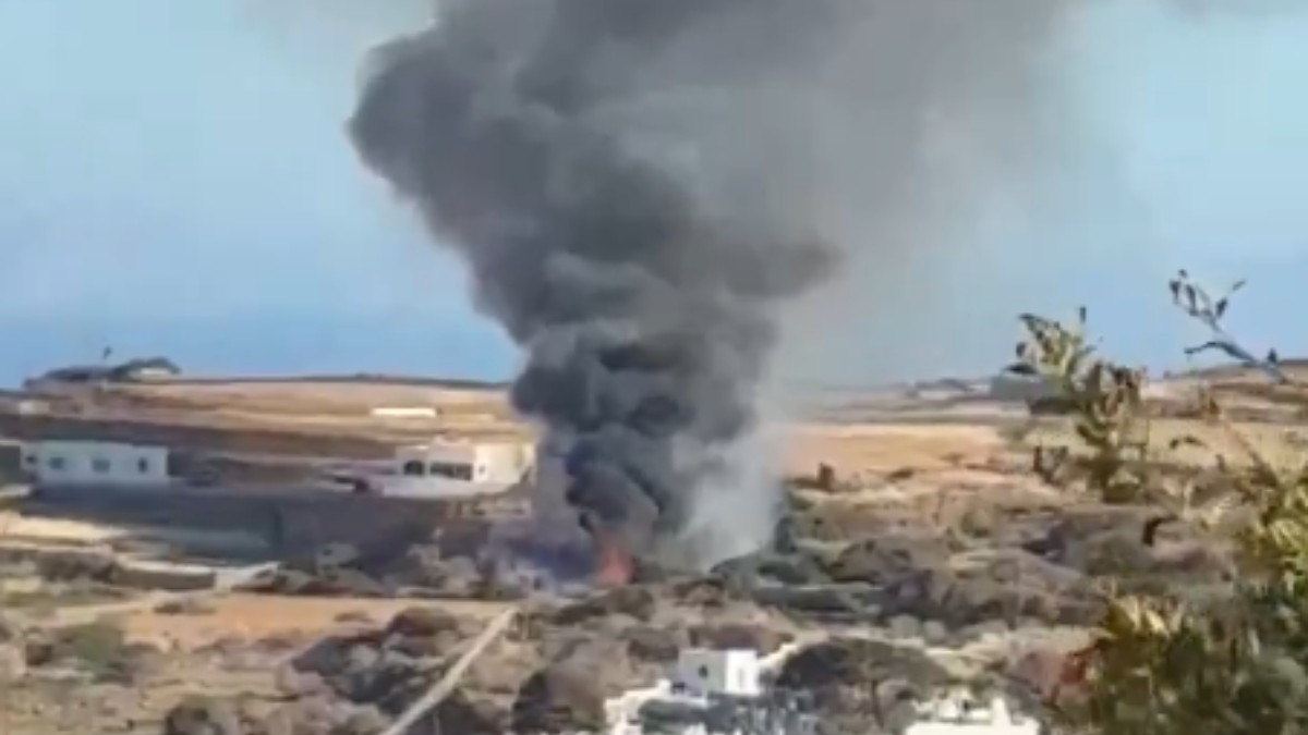 În imagine se vede o coloană densă de fum negru care se ridică de la locul unui incendiu, în apropierea unor clădiri. În jur se observă un peisaj arid, cu vegetație și teren accidentat.
