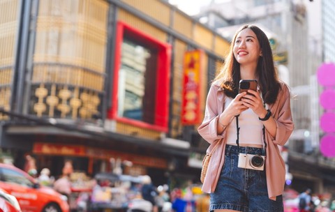 O femeie asiatică tânără cu un telefon în mână, pe o stradă din Hong Kong, în timp ce face o fotografie în fața unei clădiri înalte