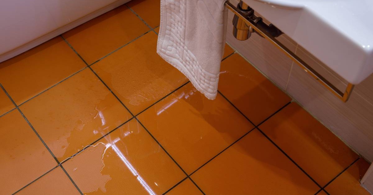 Cadru strans cu apa inundand o baie pe o gresie maru, se vede un prosop alb de hotel, agatat de o chiuveta