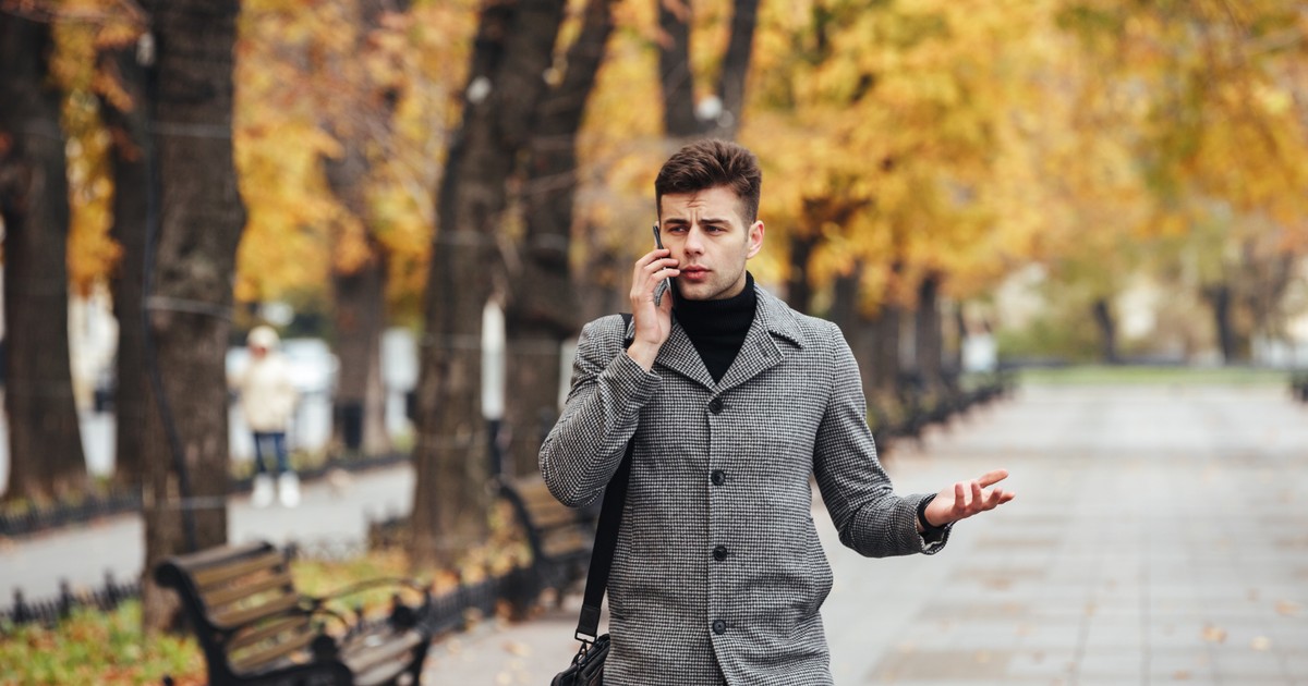 Picture of elegant male in coat with bag walking in city park and talking on smartphone in autumn