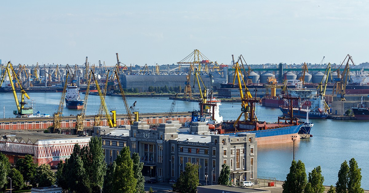 perspectivă amplă de la înălțime asupra portului Constanța, surprinzând un peisaj industrial complex și aglomerat. În prim-plan se distinge o clădire administrativă masivă, cu o arhitectură clasică, ferestre numeroase și mai multe etaje, înconjurată la bază de grupuri de copaci înalți și de câteva construcții anexe mai mici. Imediat în spatele acestei clădiri se întinde un bazin portuar în care este ancorată o navă comercială de mari dimensiuni. Dincolo de aceasta, întregul orizont este dominat de zeci de macarale înalte, cu brațe lungi și articulate, dispuse de-a lungul cheiurilor. În planul îndepărtat se conturează alte ambarcațiuni de marfă, hale industriale și o grupare de silozuri cilindrice voluminoase destinate depozitării, sugerând o infrastructură masivă și o activitate de încărcare-descărcare intensă.