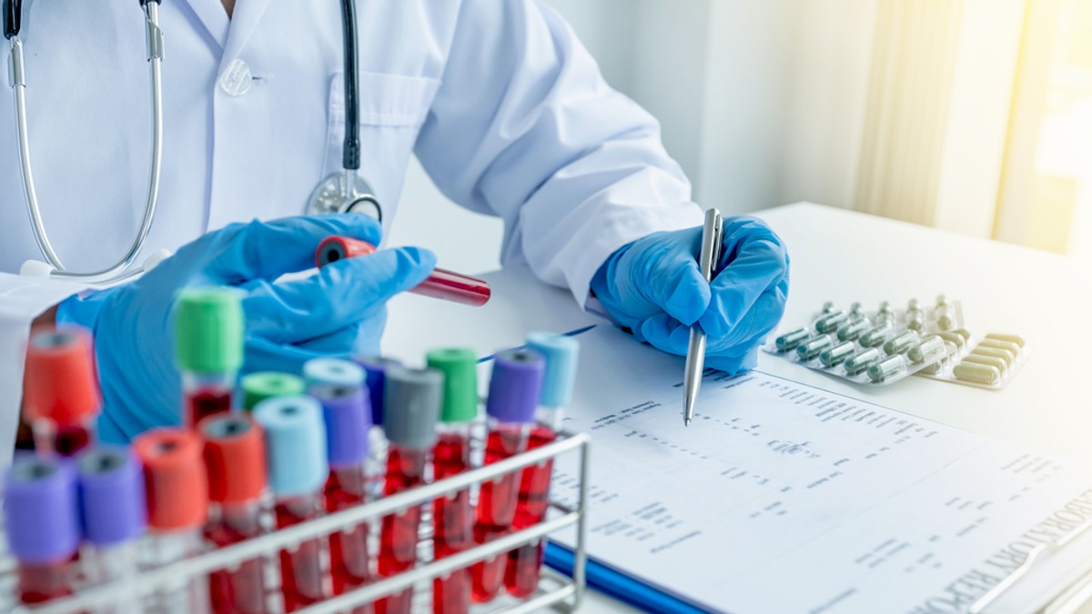 Care este diferența dintre consult și control medical - Imagine cu un medic care notează într-un tabel date de pe flacoanele de sânge pe care le are pe birou, alături de câteva folii de medicamente.