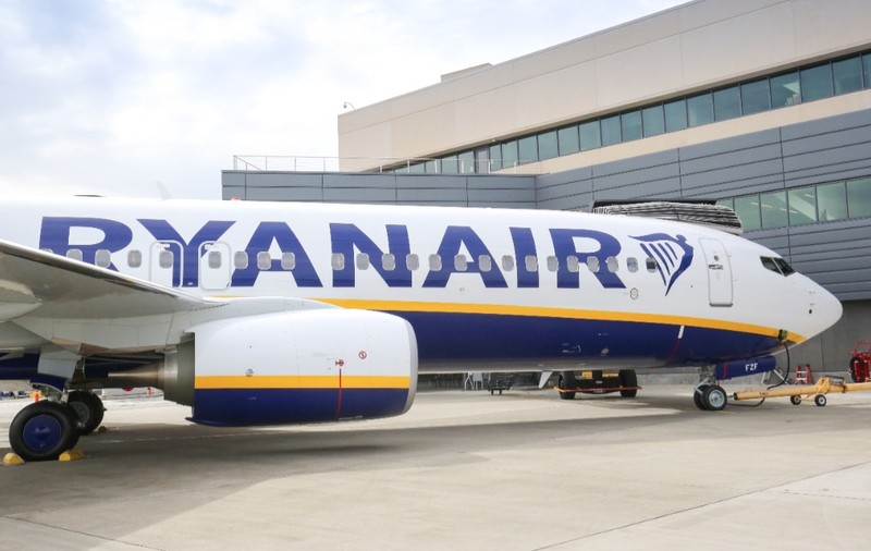 Un avion de pasageri al companiei Ryanair staționat pe pista unui aeroport, în fața unei clădiri moderne de terminal.