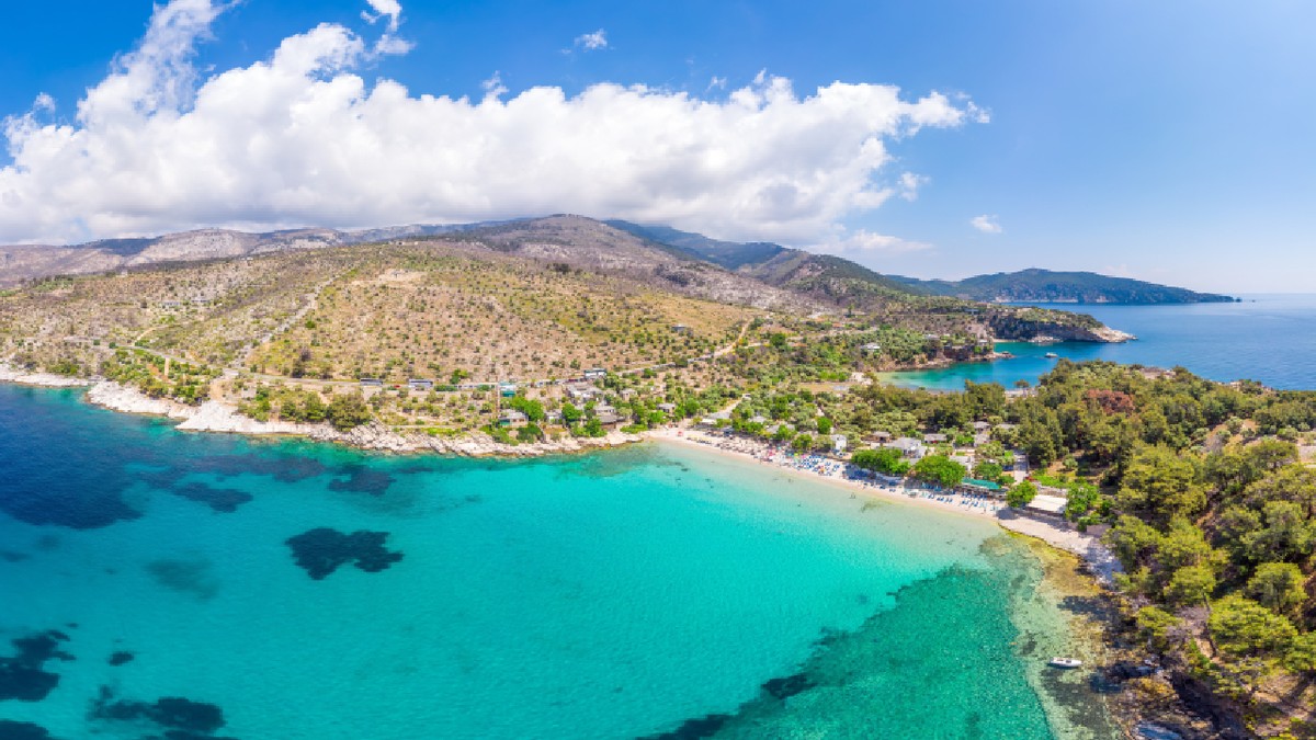 Vedere aeriană a plajei Aliki din Thassos, Grecia, cu ape de culoare turcoaz, dealuri acoperite de pini și un peisaj mediteranean liniștit,