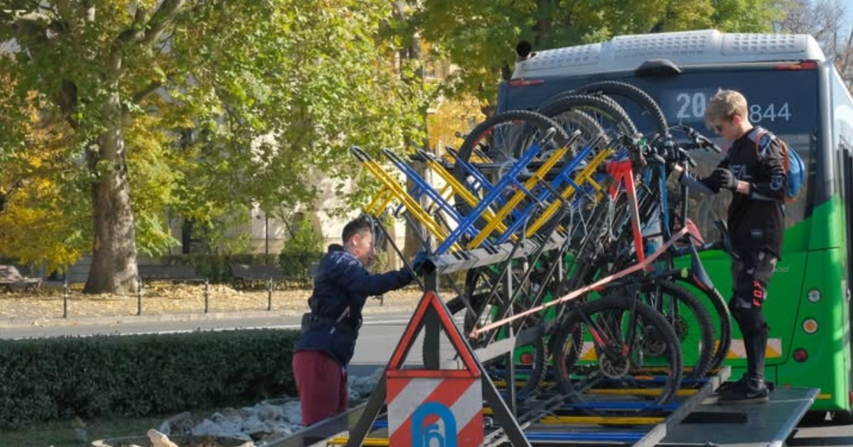 În imagine se observă un sistem de transport al bicicletelor montat în spatele unui autobuz, într-un peisaj urban. Două persoane, îmbrăcate în echipament sportiv, sunt ocupate cu încărcarea sau descărcarea bicicletelor de pe suportul special.