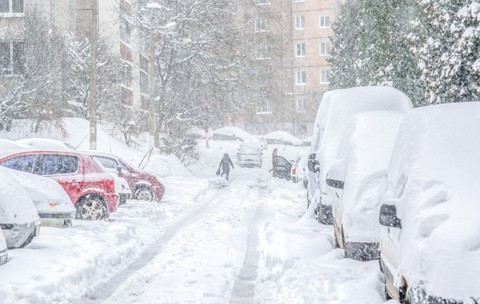O stradă dintre blocuri acoperită de un strat gros de zăpadă în timpul unei ninsori abundente, cu numeroase autoturisme parcate pe margine și o persoană care merge pe aleea îngustă curățată.