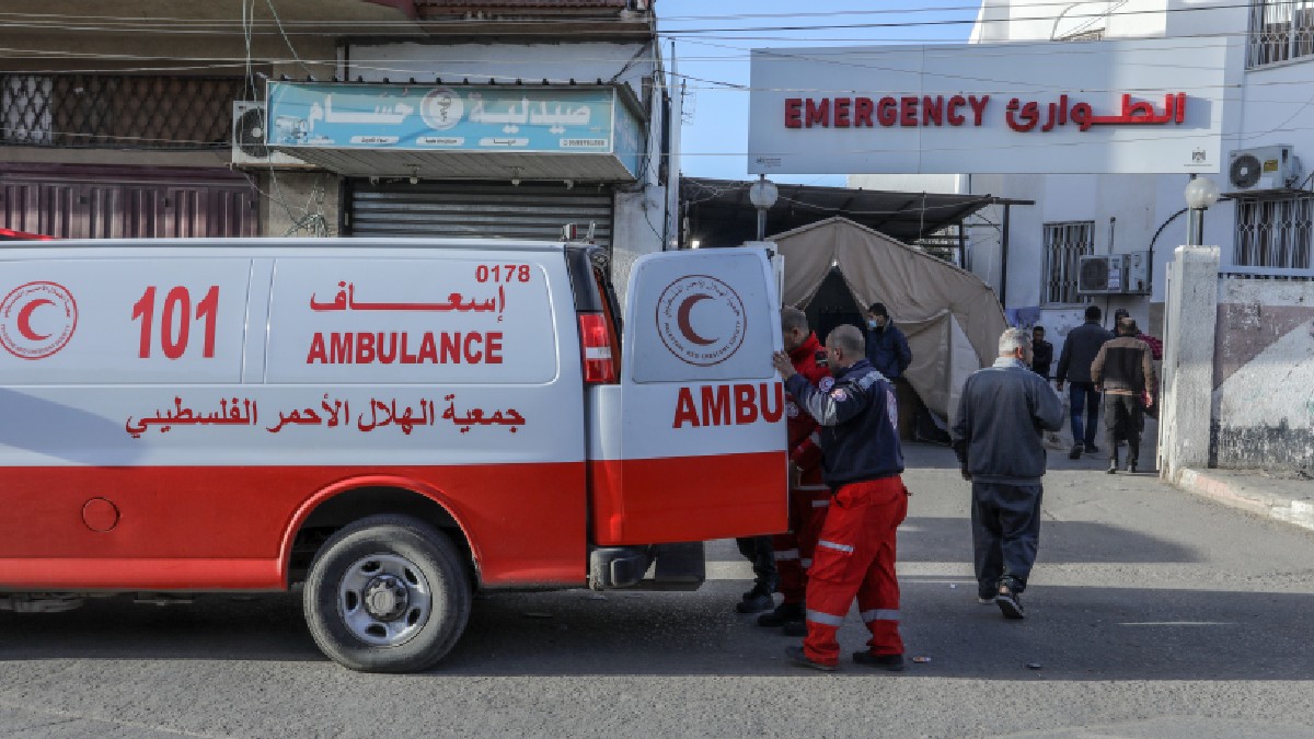 Imaginea surprinde intrarea unui spital din Fâșia Gaza, cu o ambulanță parcată în prim-plan. Ambulanța este de culoare roșu și alb, având inscripția „AMBULANCE” în engleză și „إِسْعَاف” în arabă. De asemenea, pe vehicul se află sigla Semilunii Roșii, alături de textul „جمعیة الهلال الأحمر الفلسطيني” (Societatea Semilunii Roșii Palestiniene). La intrarea spitalului, deasupra unei uși, se observă un panou alb pe care scrie „EMERGENCY” și „الطوارئ” în arabă. În curtea spitalului se văd mai multe corturi, sugerând că este un punct de triaj sau un spațiu de urgență improvizat. Mai multe persoane, inclusiv personal medical, se află în zona de intrare. Atmosfera generală a imaginii este tensionată și sugerează un mediu de criză, specific unei zone de conflict.