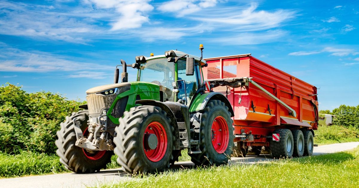 Un tractor cu remorcă de încărcare pe un drum, în timpul recoltării. Pe jos este iarbă verde și se vede un cer senin