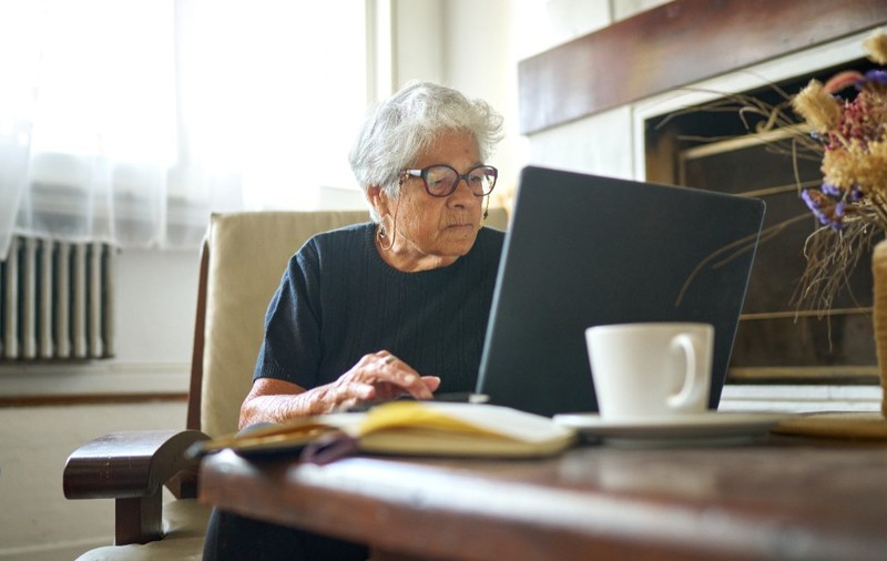 O pensionară cu părul cărut, în timp ce stă la un birou din sufragerie, în fața unui laptop și plătește facturi