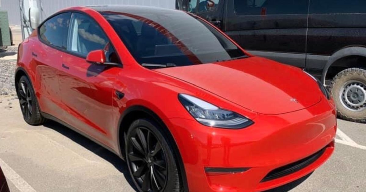 O mașină Tesla roșie, într-o parcare, alături de un microbuz negru