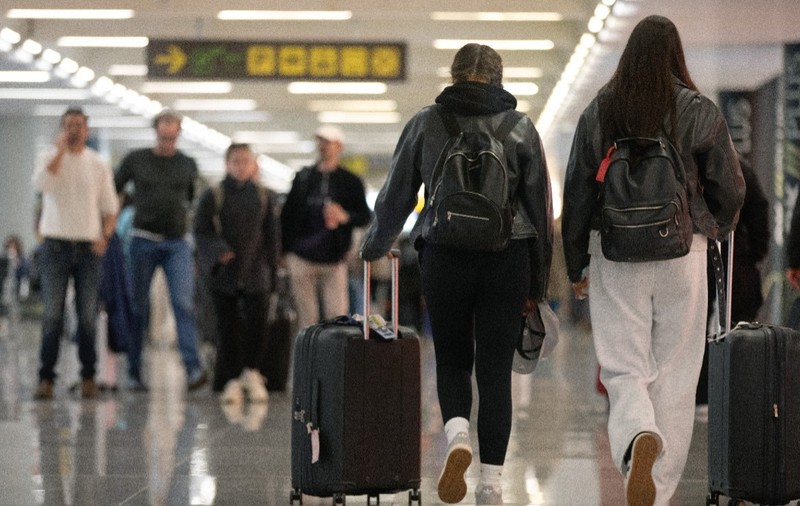 Două persoane văzute din spate mergând printr-un terminal de aeroport și trăgând după ele valize cu rotile, într-o zonă tranzitată și de alți pasageri.