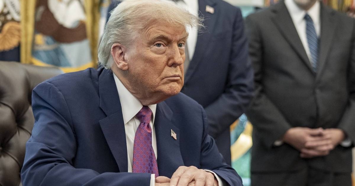 Președintele american Donald Trump în timp ce stă la biroul său de la Casa Albă, are mâinile încrucișate pe birou, poartă un costum albastru și o cravată mov.