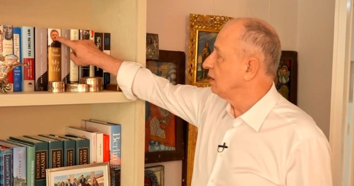 Mircea Geoană, îmbrăcat cu o cămașă albă, în timp ce arată cu degetul către o carte din biblioteca sa