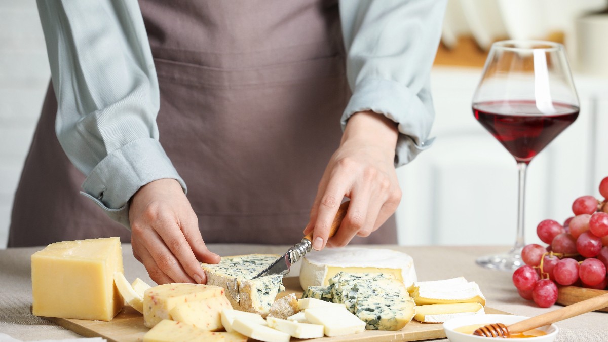 Imaginea arată o persoană care pregătește un platou cu diverse brânzeturi, inclusiv sortimente cu mucegai albastru, brânzeturi moi și tipuri maturate. Pe masă se află și un pahar de vin roșu, struguri și un bol cu miere, sugerând un aranjament rafinat, ideal pentru o ocazie festivă precum masa de Crăciun.