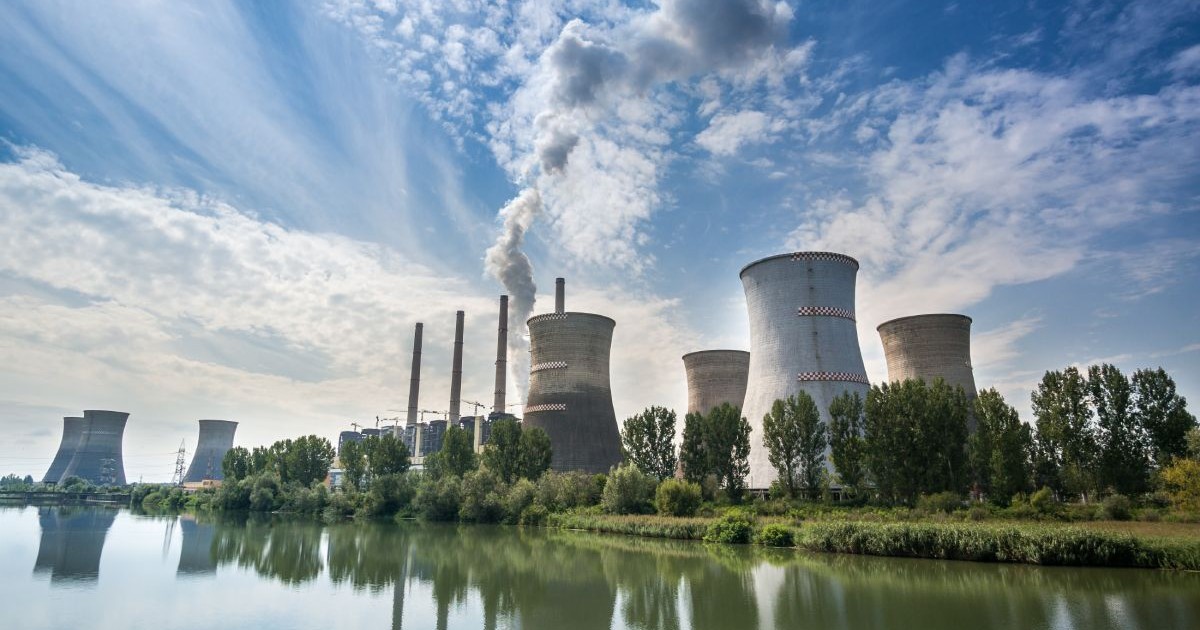 Termocentrala Turceni, cu un lac în față, turnurile uriașe pe fundal pe care iese fum și cerul albastru cu nori albi