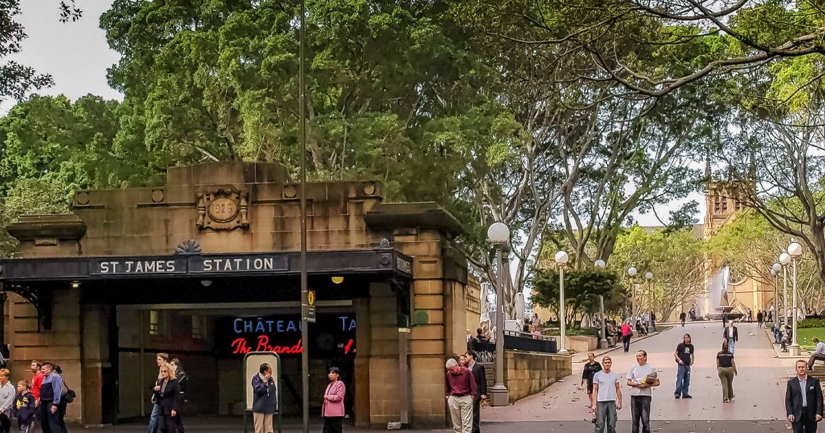 Stația istorica de metrou St James din Hyde Park, Sydney, Australia, se vad mai multi oameni, intrarea la metrou cu un parc si copaci mari, inverziti
