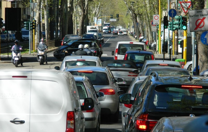 Trafic aglomerat pe un bulevard urban mărginit de copaci, cu numeroase mașini și scutere oprite sau deplasându-se în coloană lângă un semafor și indicatoare rutiere.