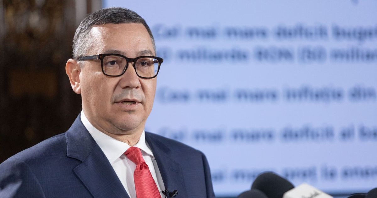 Victor Ponta, îmbrăcat în costum cu sacou de culoare albastru închis, cămașă albă și cravată roșie, iar pe fundal un panou pe care sunt cuvinte în blur