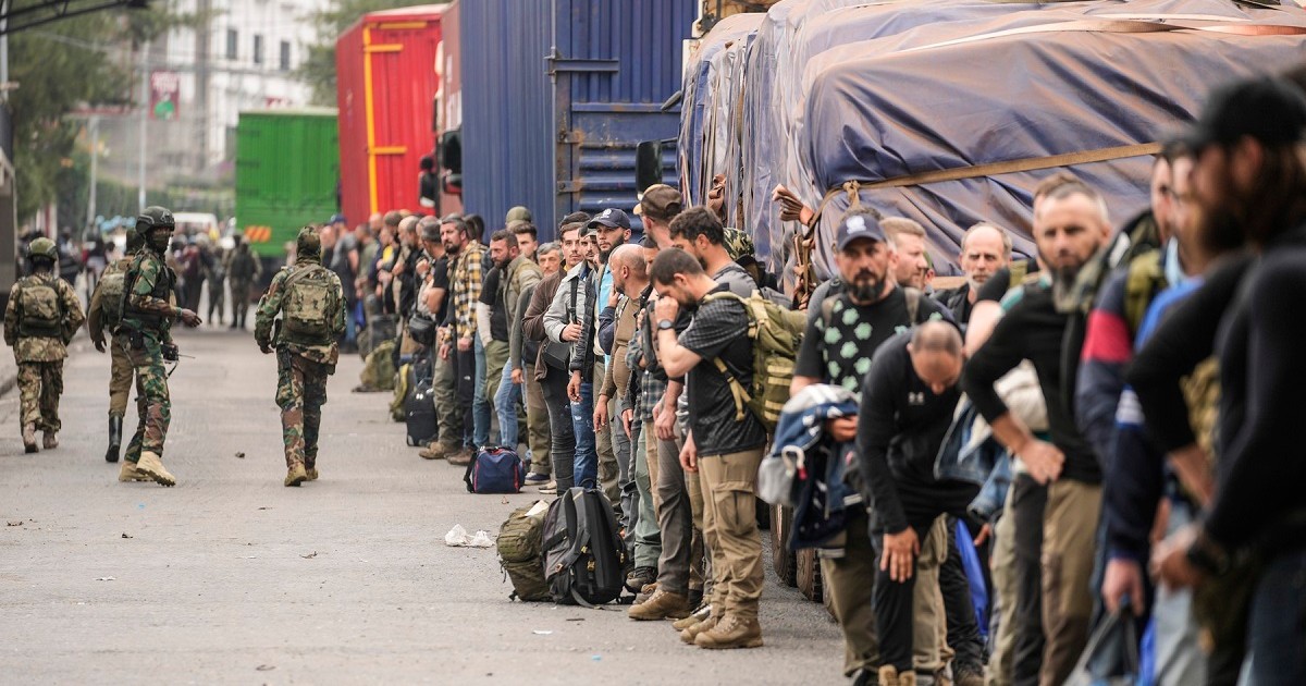 Mai mulți bărbați sunt aliniați în șir indian lângă vehicule și sunt păziți de militari