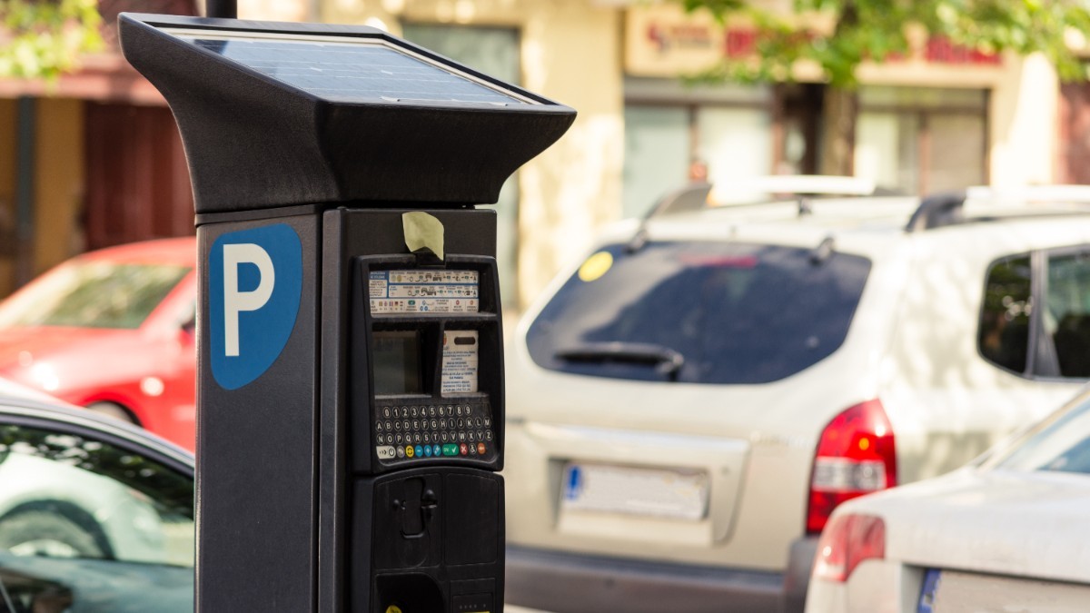 Prim-plan cu un aparat electronic de plată a parcării, alimentat cu panou solar, amplasat pe marginea unei străzi cu mașini parcate.