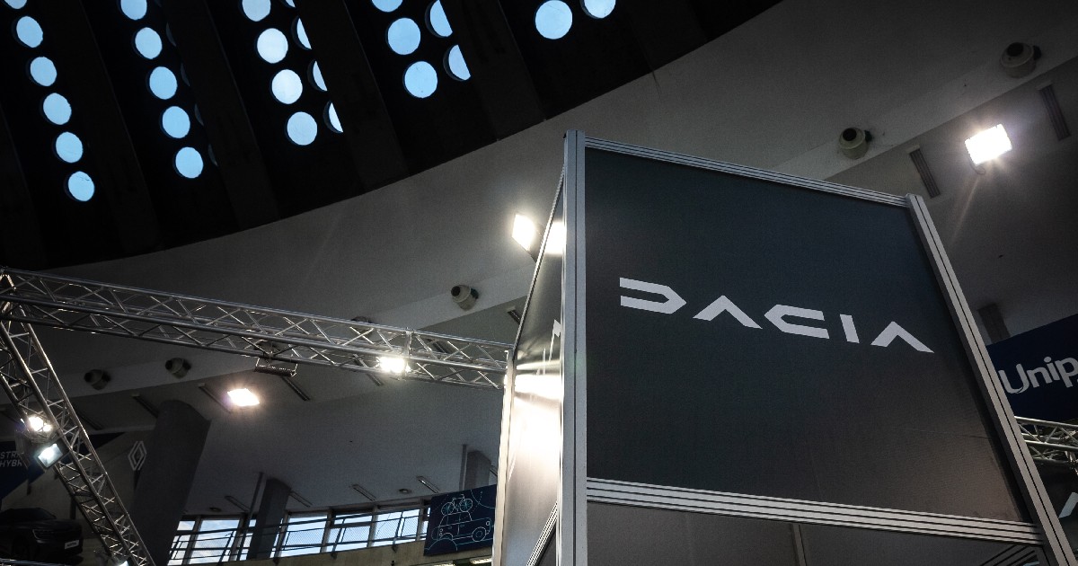 Imaginea surprinde un stand al mărcii de mașini Dacia, probabil la un târg sau un salon auto, judecând după structura metalică a standului și luminile de spot. Pe un panou mare, de culoare închisă, este scris cuvântul "DACIA" cu litere albe, stilizate. Deasupra standului se observă o structură arhitecturală a tavanului, compusă dintr-un rând de ferestre circulare, iluminate, ce lasă să pătrundă o lumină difuză, dar și structuri metalice de susținere și reflectoare, specifice unui spațiu expozițional.
