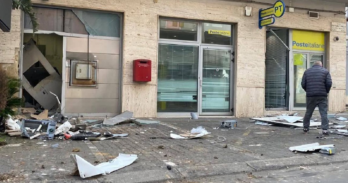 O fotografie surprinde o scenă haotică pe stradă, în fața unei clădiri. Resturi și moloz acoperă solul, inclusiv sticlă spartă, lemn și metal. Un bărbat îmbrăcat într-o jachetă închisă la culoare stă în picioare în dreapta. Un copac ascunde parțial partea stângă a imaginii.