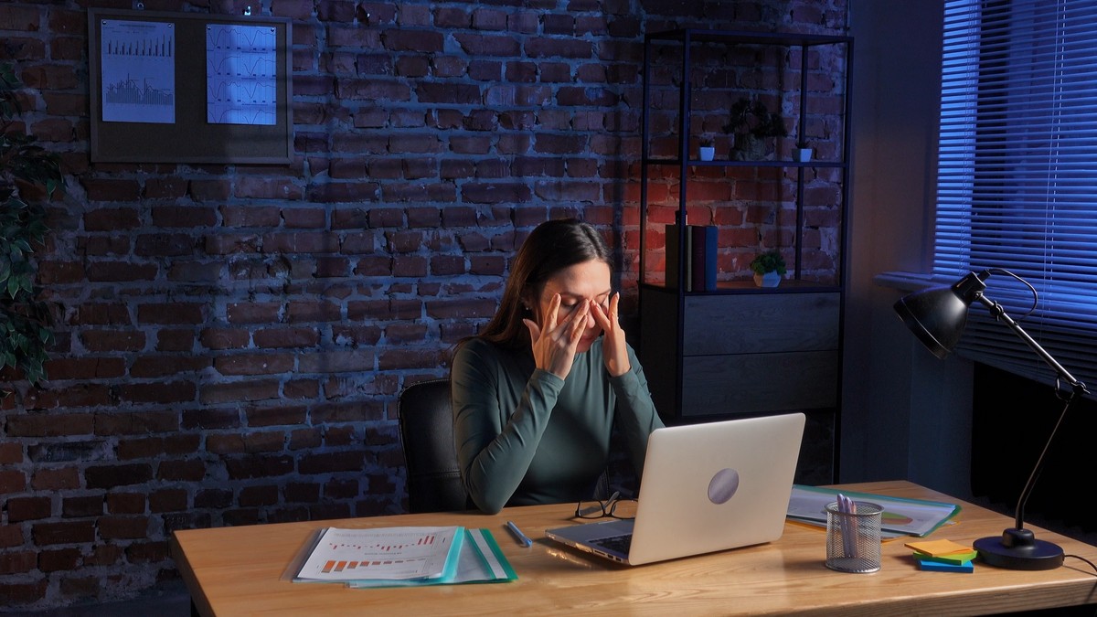 Femeie care lucreaza la laptop. Se afla la birou, in jur are rapoarte si pare ca o doare capul