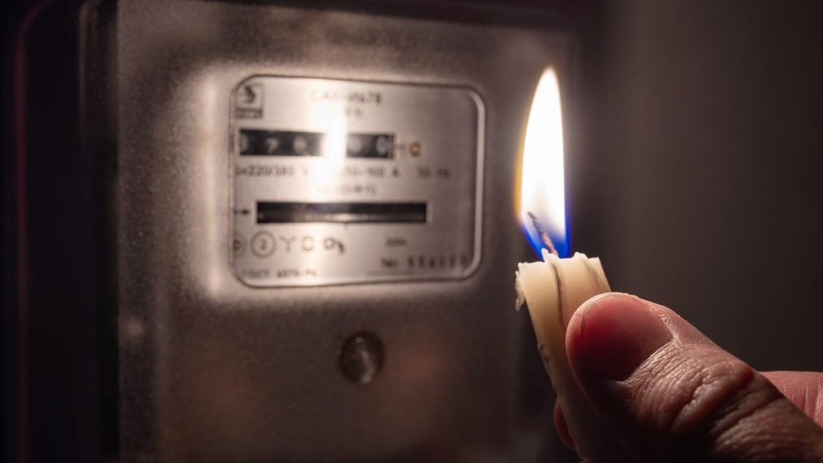 Mâna unui bărbat care ține o lumânare aprinsă și se uită la contorul de electricitate din casă, într-o cameră aflată în întuneric