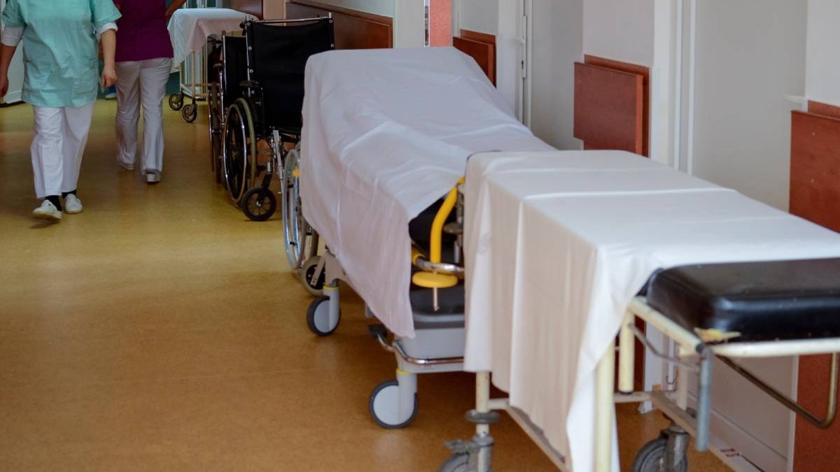 Hol de spital, cu doua paturi in dreapta acoperite cu cearșafuri albe, in stanga se vad picioarele unor cadre medicale
