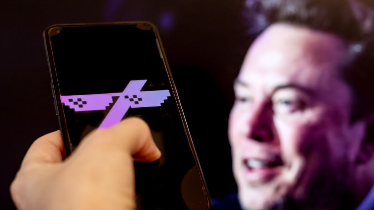 sigla aplicației Grok este afișată pe un smartphone, în timp ce în fundal, pe ecranul unui computer, se vede un portret al lui Elon Musk