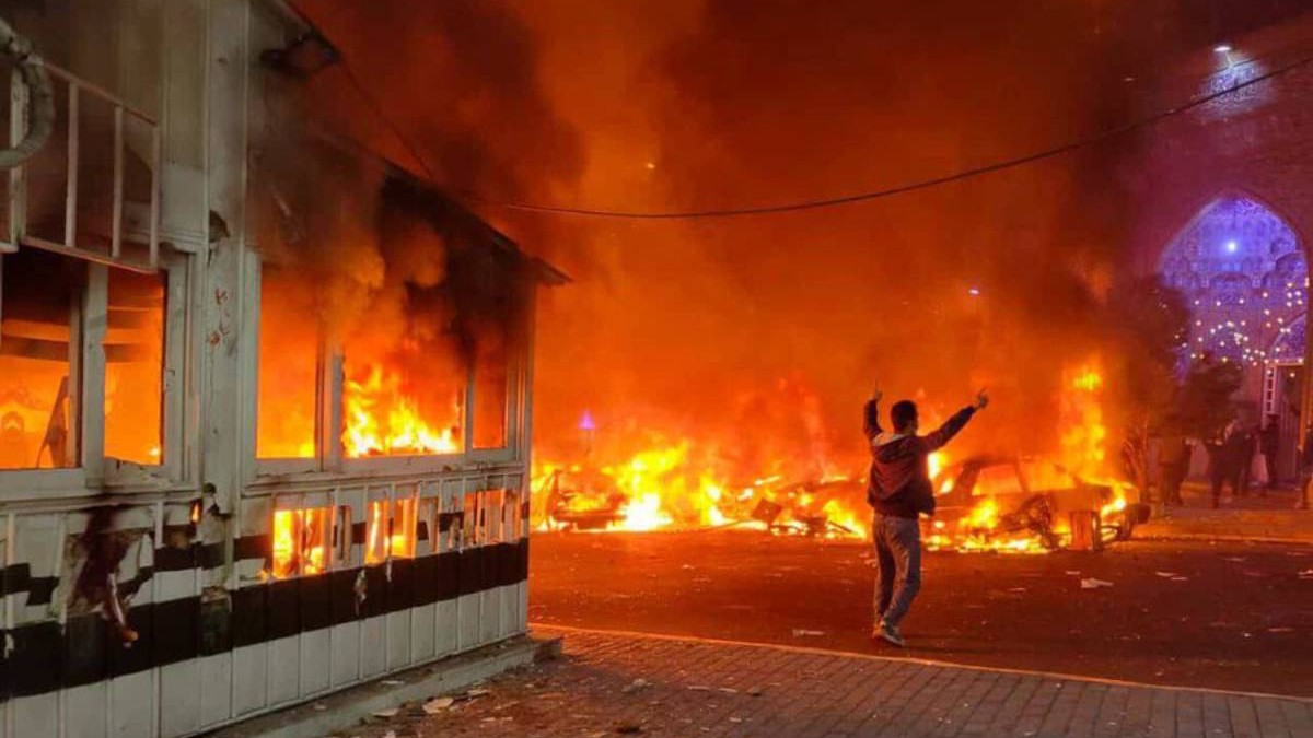 Imagine de noapte din timpul protestelor din Iran, se vad flacari mari in intuneric, care au cuprins totul, silueta unei persoane cu mainile ridicate in mijlocul lor