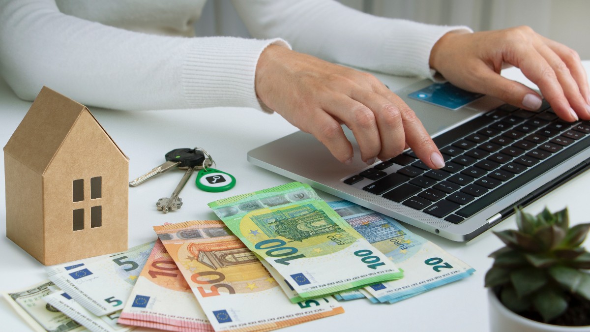 O femeie la birou care scrie pe tastatură. Pe birou se află bancnote de euro și o casă în miniatură