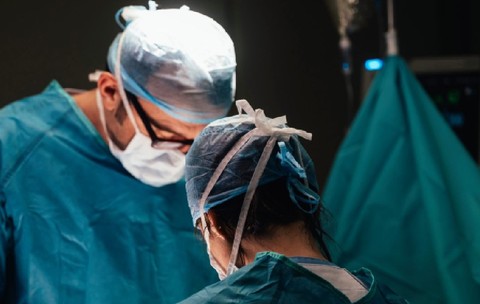 Doi medici, costumati in halate cu bonete si masti, care opereaza in sala de operatie