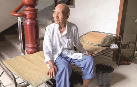 Un bărbat de 100 de ani din China stă pe un pat pliant, poarta o bluza alba si pantaloni albastri