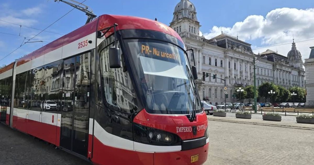 Tramvai de culoare roșie, în centrul municipiului Arad, cu cerul albastru pe fundal și o clădire istorică