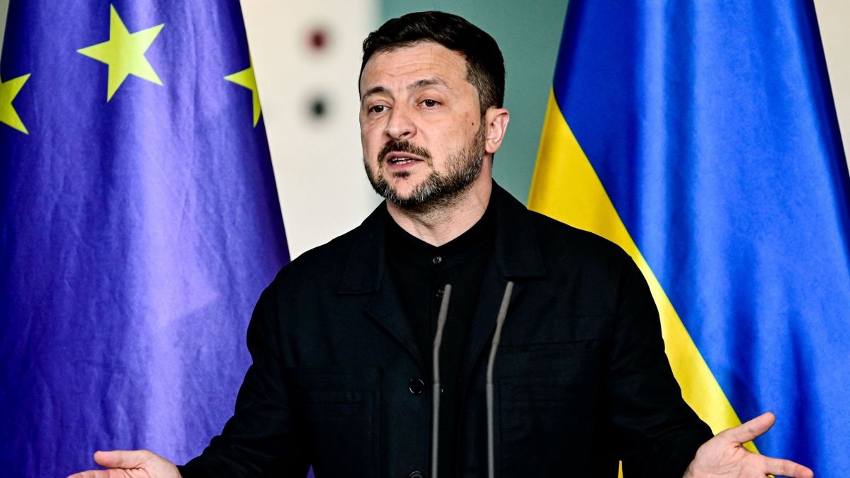 Volodimir Zelenski, îmbrăcat într-o cămașă neagră, dă explicații cu mâinile larg deschise în timpul unei conferințe de presă, având în spate steagurile Ucrainei și al Uniunii Europene