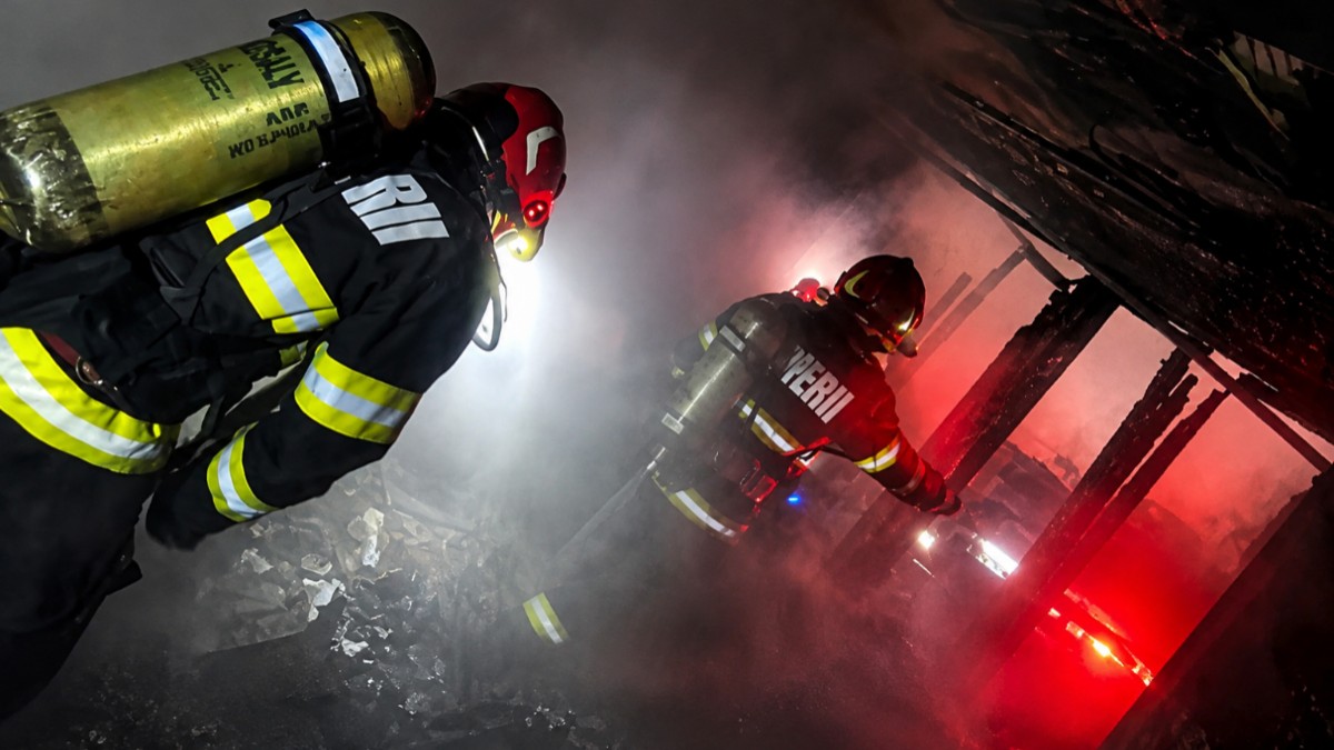 Imaginea surprinde pompieri în costume de protecție și tuburi de oxigen care intervin într-un spațiu plin de fum și flăcări pentru stingerea unui incendiu.