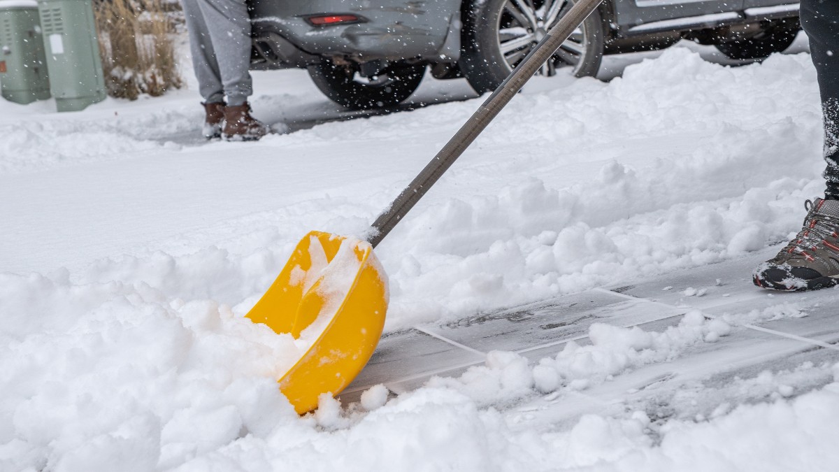 O lopată galbenă îndepărtează zăpada proaspăt căzută de pe un trotuar, în timp ce fulgii continuă să cadă. În fundal se văd mașini parcate și persoane îmbrăcate gros, semn că deszăpezirea are loc în plină iarnă.